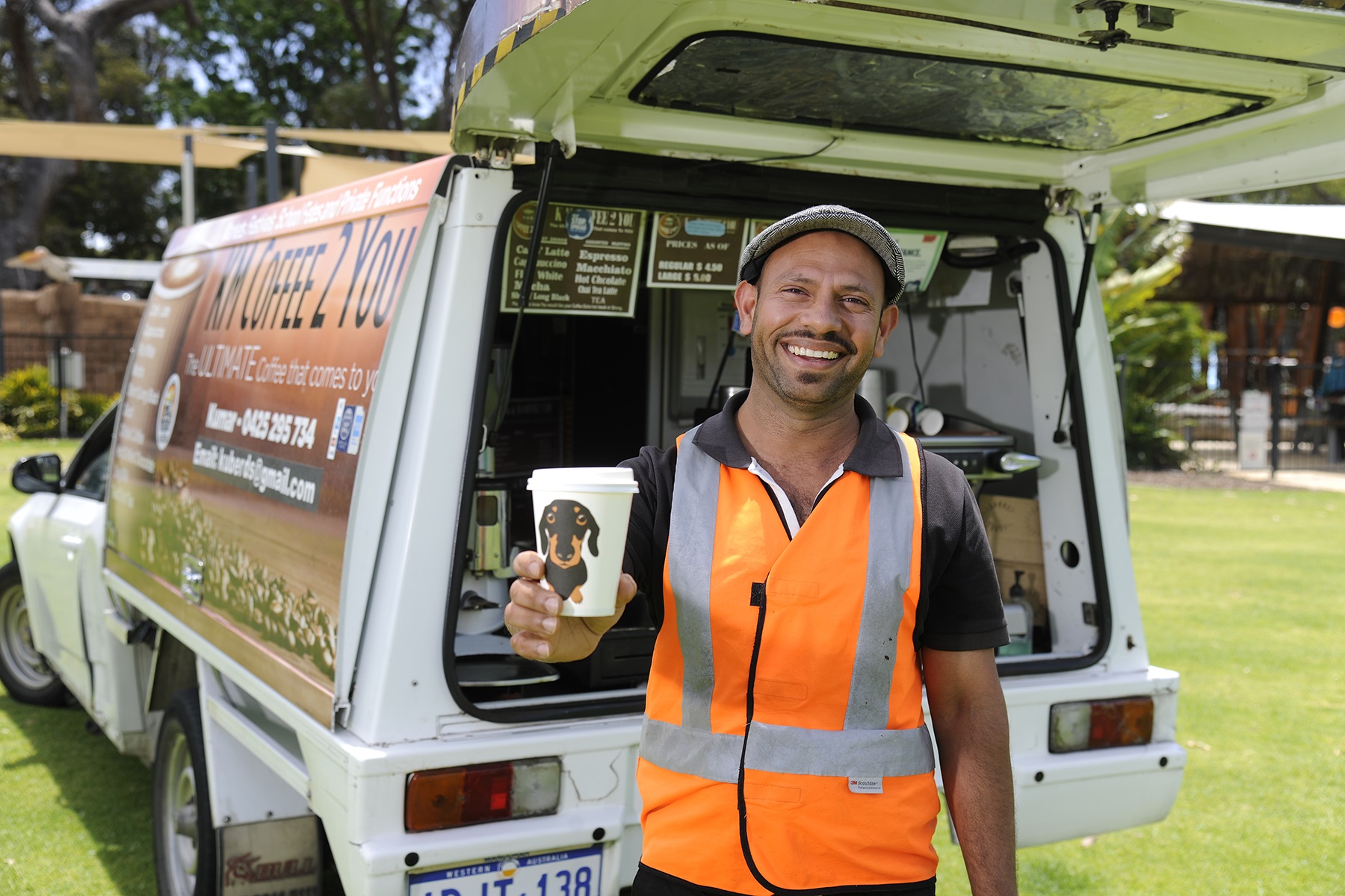 Photo of a man standing in front of a coffee van. He is smiling at the camera and holding up a take away cup of coffee.